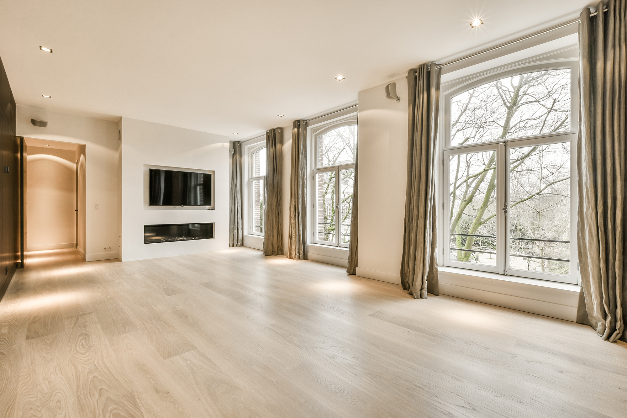 an empty living room with wood flooring and large windows looking out onto the trees that line the street outside