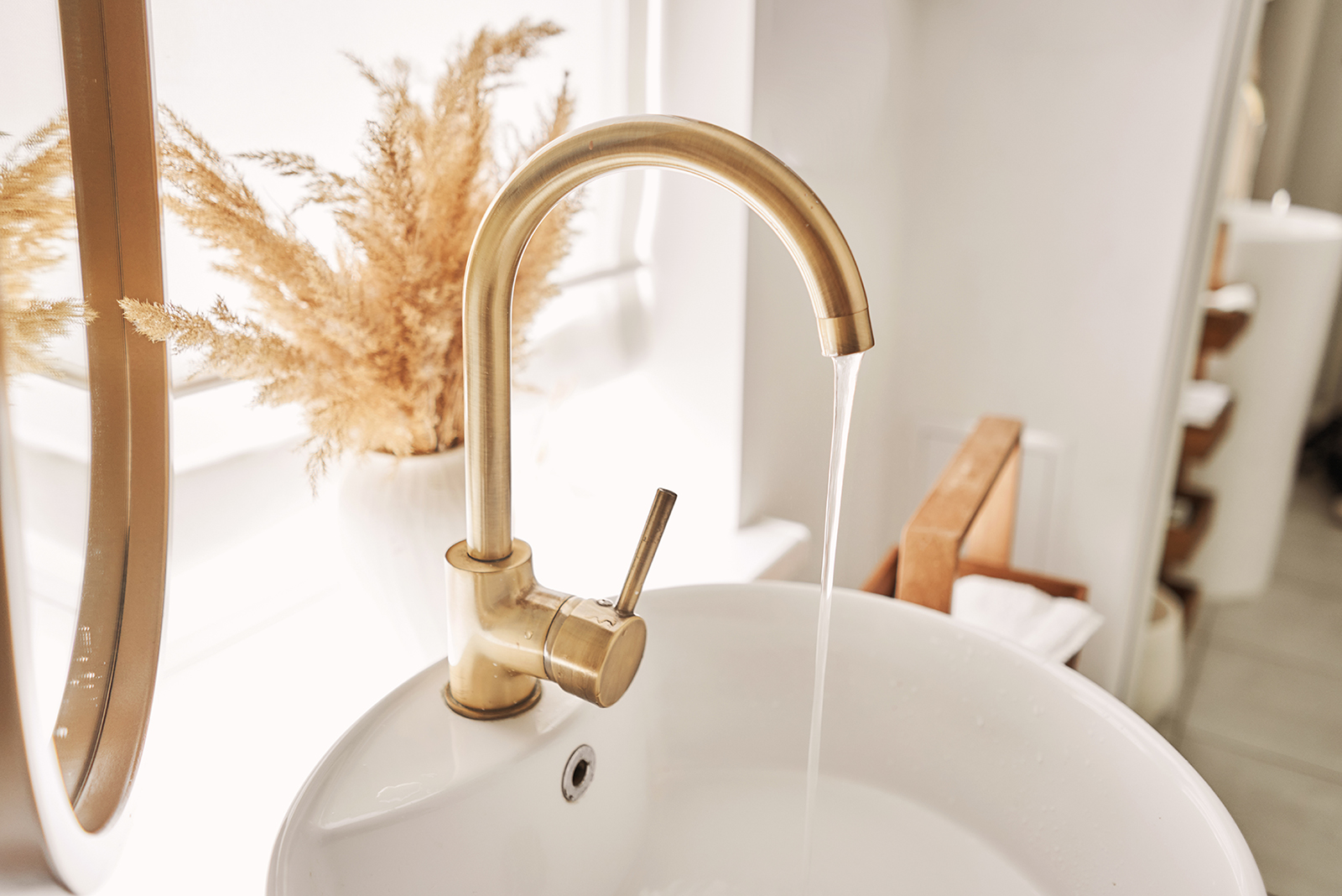 Close-up of an elegant golden faucet in the bathroom sink next to stylish decorations. A beautiful sink with a golden faucet next to an oval mirror and a shelf with hand towels.